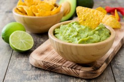 Guacamole zubereiten: eine Holzschale mit Guacamole, darin ein Tortilla-Chip, daneben eine Schale mit mehr Chips und eine halbierte Limette.