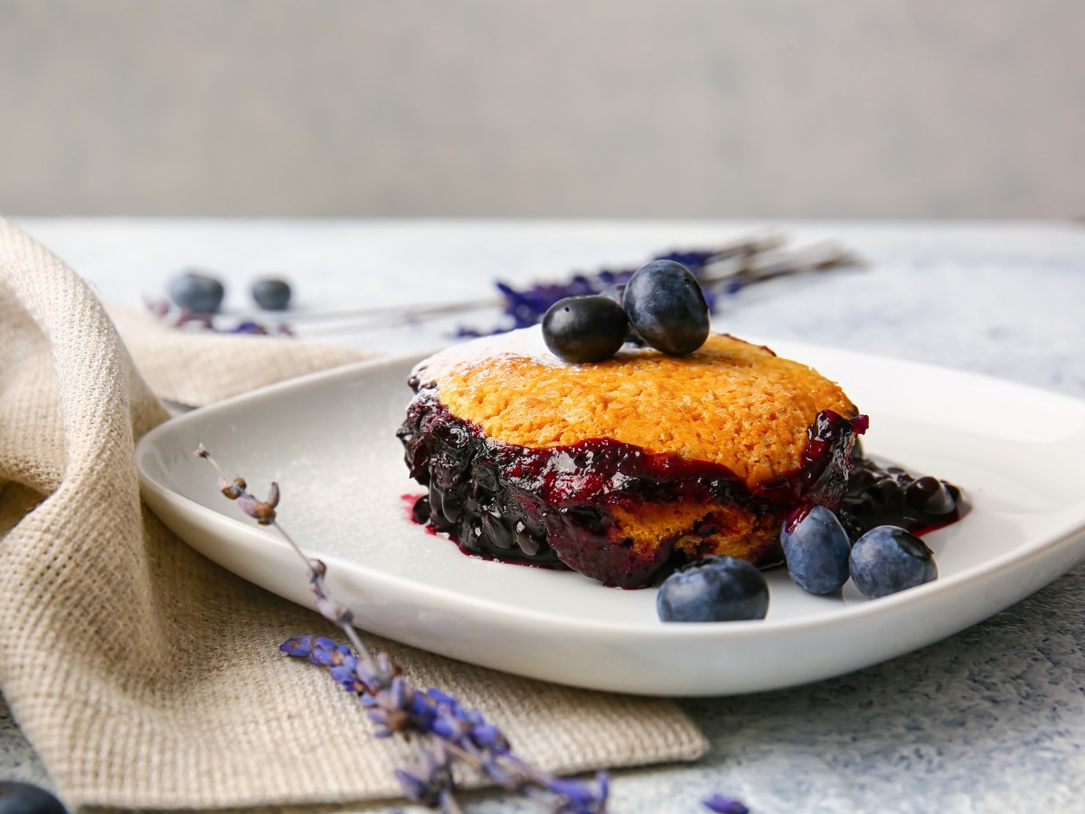 Eine Portion Heidelbeer-Auflauf mit frischen Blaubeeren auf einem Teller, daneben drapiert eine Stoffserviette und Blüten.