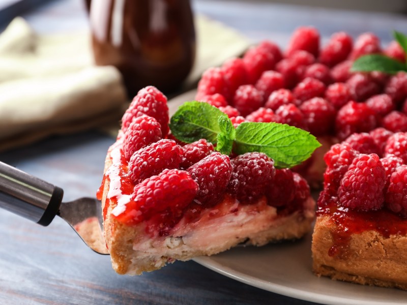 Himbeer-Schmand-Kuchen, aus dem ein Stück mit einem Tortenheber herausgenommen wird.