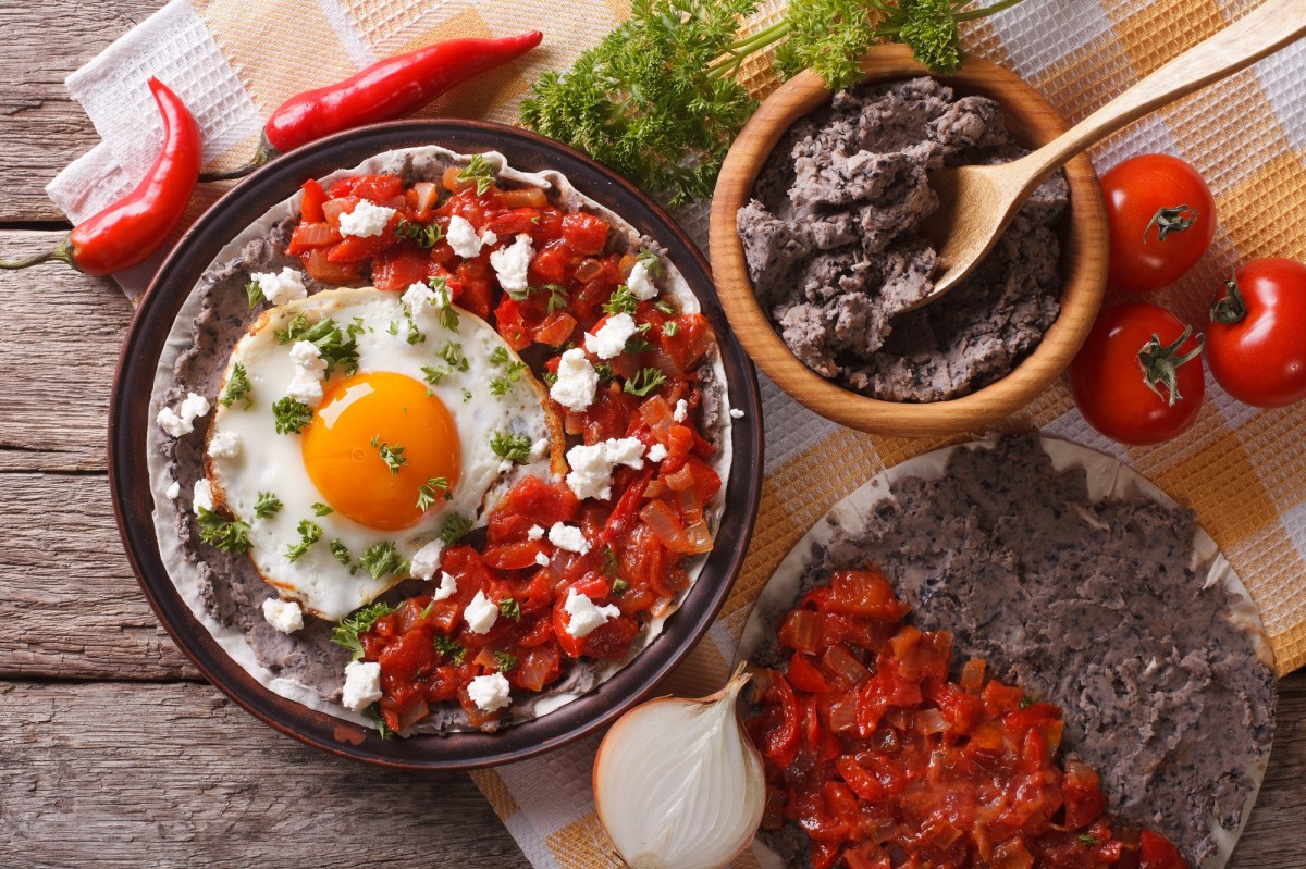 Huevos Rancheros auf einem dunklen Teller. Daneben eine Schale mit Bohnenmus, eine weitere Tortilla mit Tomatensoße und Bohnenmus und Tomaten, Chilis, Zwiebeln und Petersilie. Draufsicht.