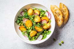 Kartoffelsalat mit Erbsen, Radieschen und Rucola in einer weißen Schüssel. Daneben geröstetes Brot.