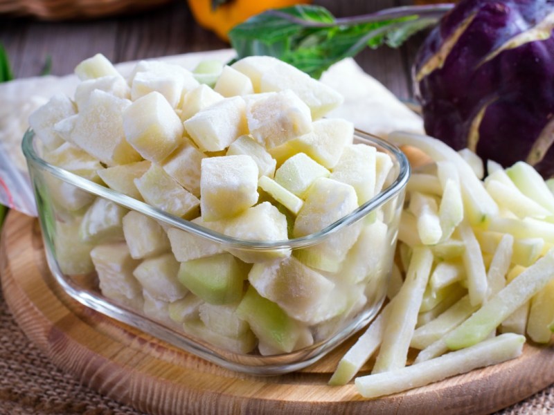 Kohlrabi einfrieren: gefrorene Kohlrabistücke in einer Glasschüssel. Im Hintergrund liegt frisches Gemüse.