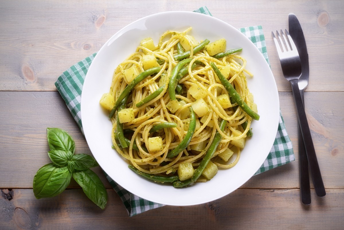 Ligurische Pasta mit Bohnen und Kartoffeln auf einem weißen Teller. Daneben liegt Besteck und etwas Basilikum.