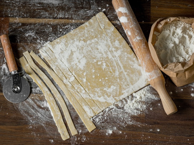 Nudelteig ohne Ei, ausgerollt und teilweise geschnitten, auf einer dunklen Holzunterlage. Daneben liegen ein Nudelholz und ein Pizzaschneider.