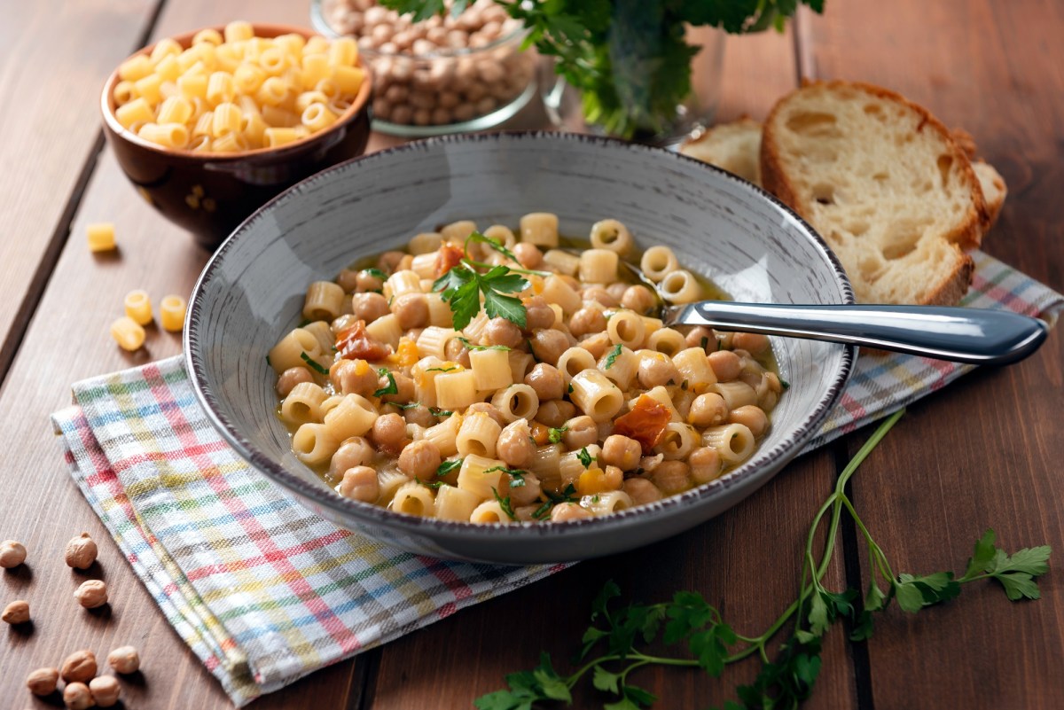 Pasta e Ceci in einer grauen Schüssel. Daneben Schalen mit Nudeln und Kichererbsen sowie Petersilie und zwei Scheiben Brot.