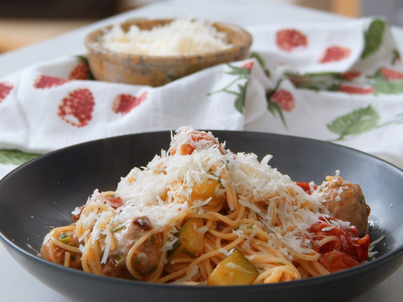 Ein schwarzer Teller mit einer Pasta-Pfanne mit Fleischbällchen, Gemüse und geriebenem Parmesan.