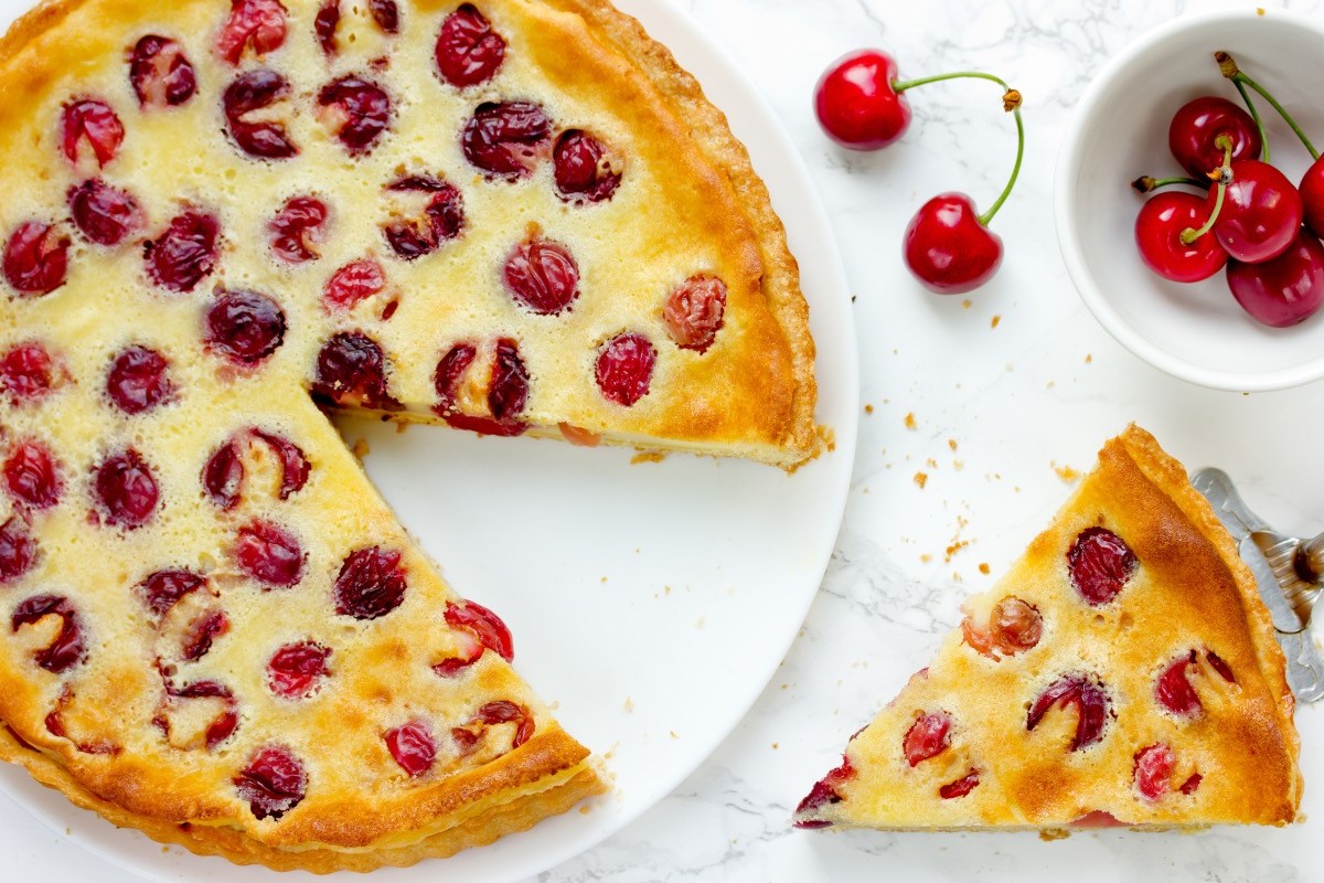 Ein Pudding-Kirschkuchen, aus dem ein Stück geschnitten wurde, in der Draufsicht. Das Kuchenstück sowie eine Schale mit frischen Kirschen sind daneben.
