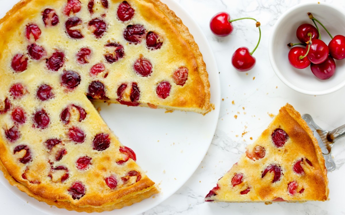 Ein Pudding-Kirschkuchen, aus dem ein Stück geschnitten wurde, in der Draufsicht. Das Kuchenstück sowie eine Schale mit frischen Kirschen sind daneben.