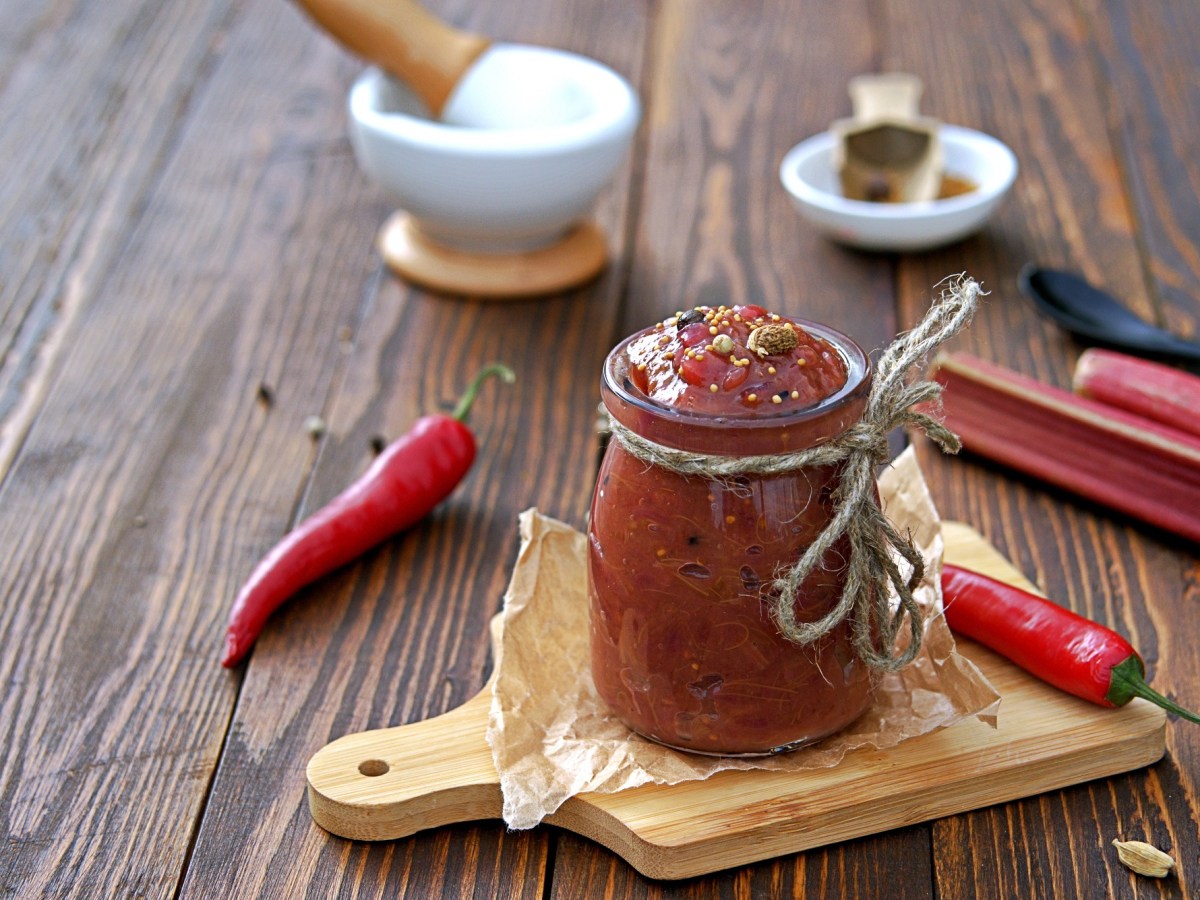 Glas mit Rhabarber-Chutney auf Brett, Chilis und Rhabarber daneben.