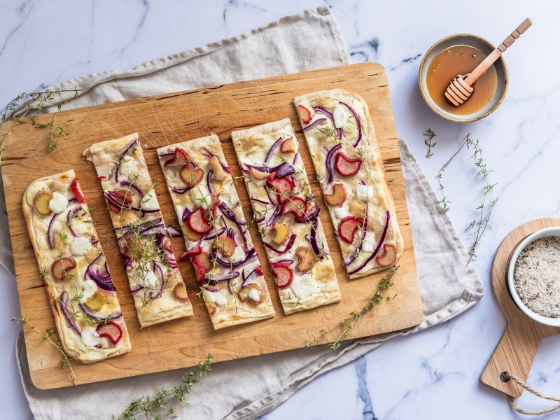 Brett mit Rhabarber-Flammkuchen auf einem hellen Untergrund mit Schale Honig und Salt daneben.