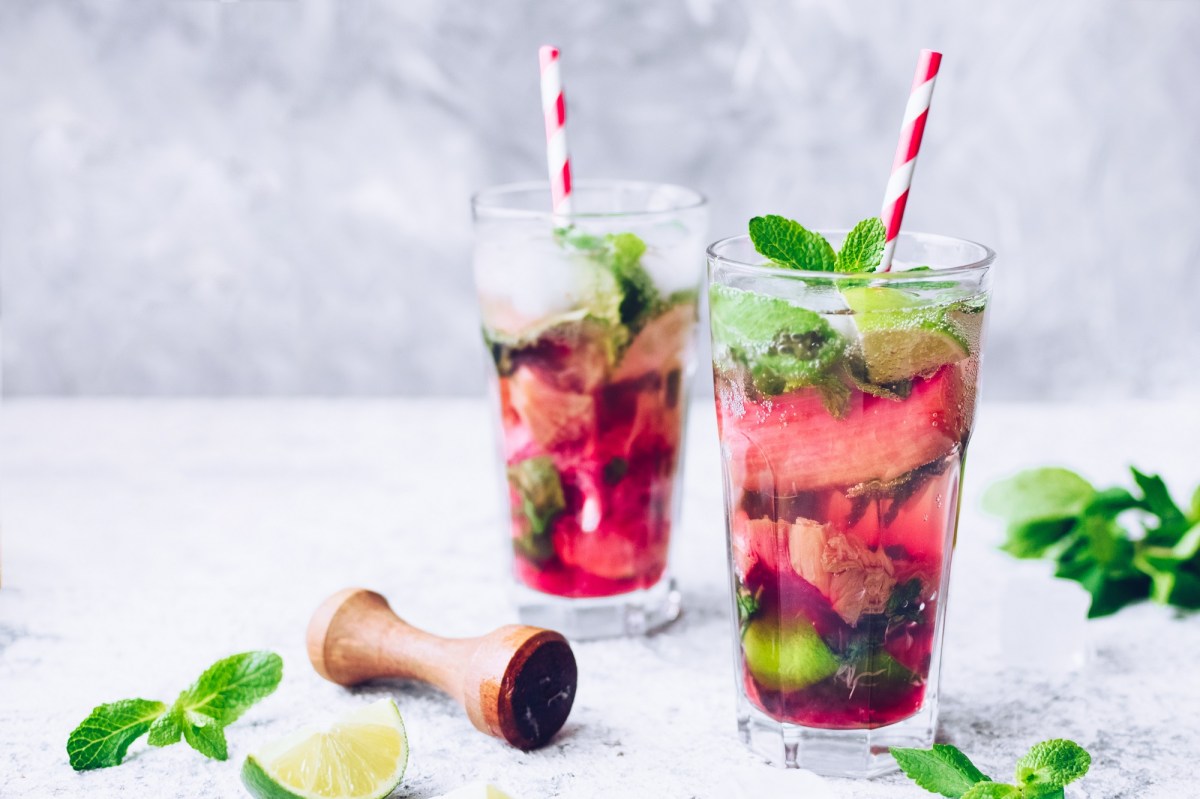 Zwei Gläser Rhabarber-Mojito mit Minze, Limette und Barstößel daneben.