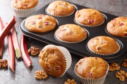 Ein Muffinblech mit Rhabarber-Walnuss-Muffins, zwei Muffins liegen daneben, außerdem Rhabarberstangen und Walnüsse daneben.