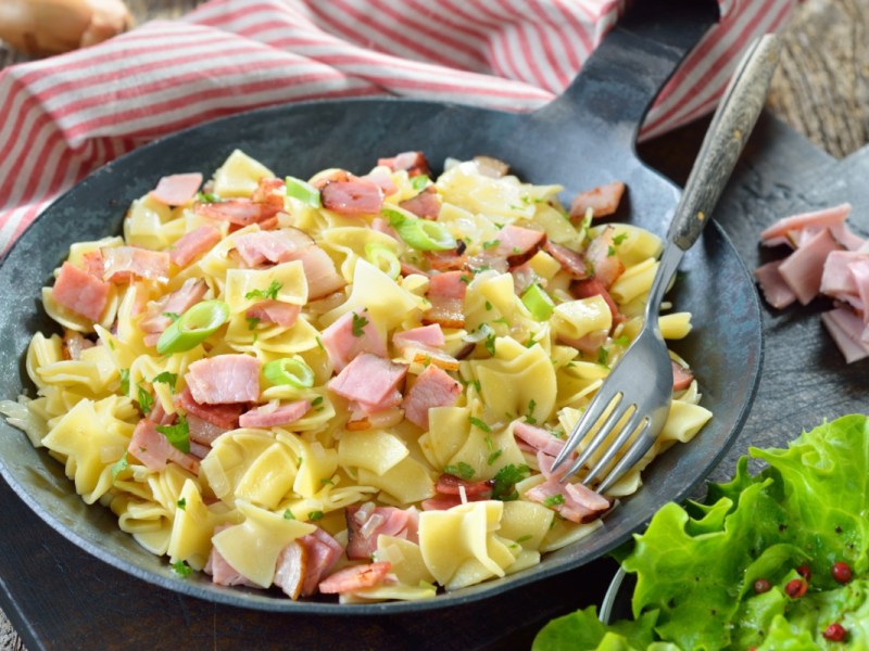 Schinkenfleckerl in einer Pfanne. Daneben ein Salat.