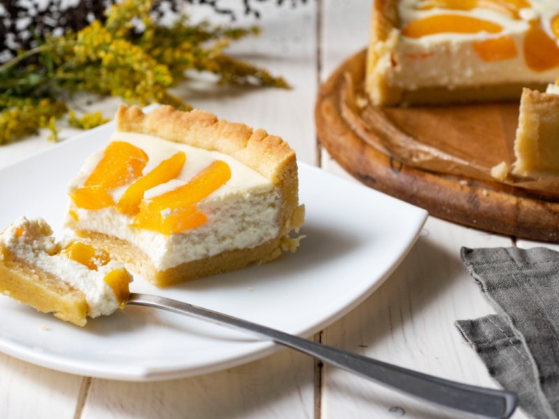 Ein Stück Schmandkuchen mit Pfirsichen auf einem Teller. Im Hintergrund steht der restliche Kuchen.