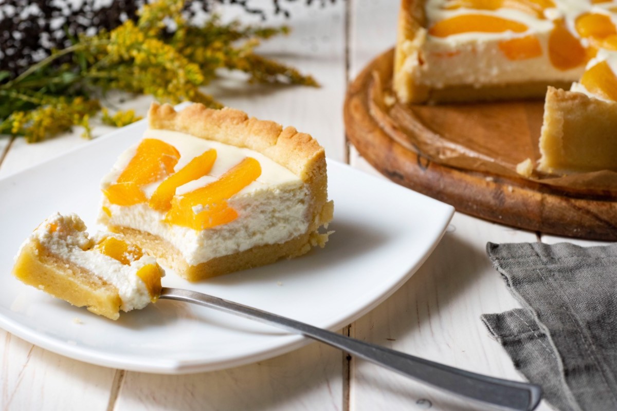 Ein Stück Schmandkuchen mit Pfirsichen auf einem Teller. Im Hintergrund steht der restliche Kuchen.