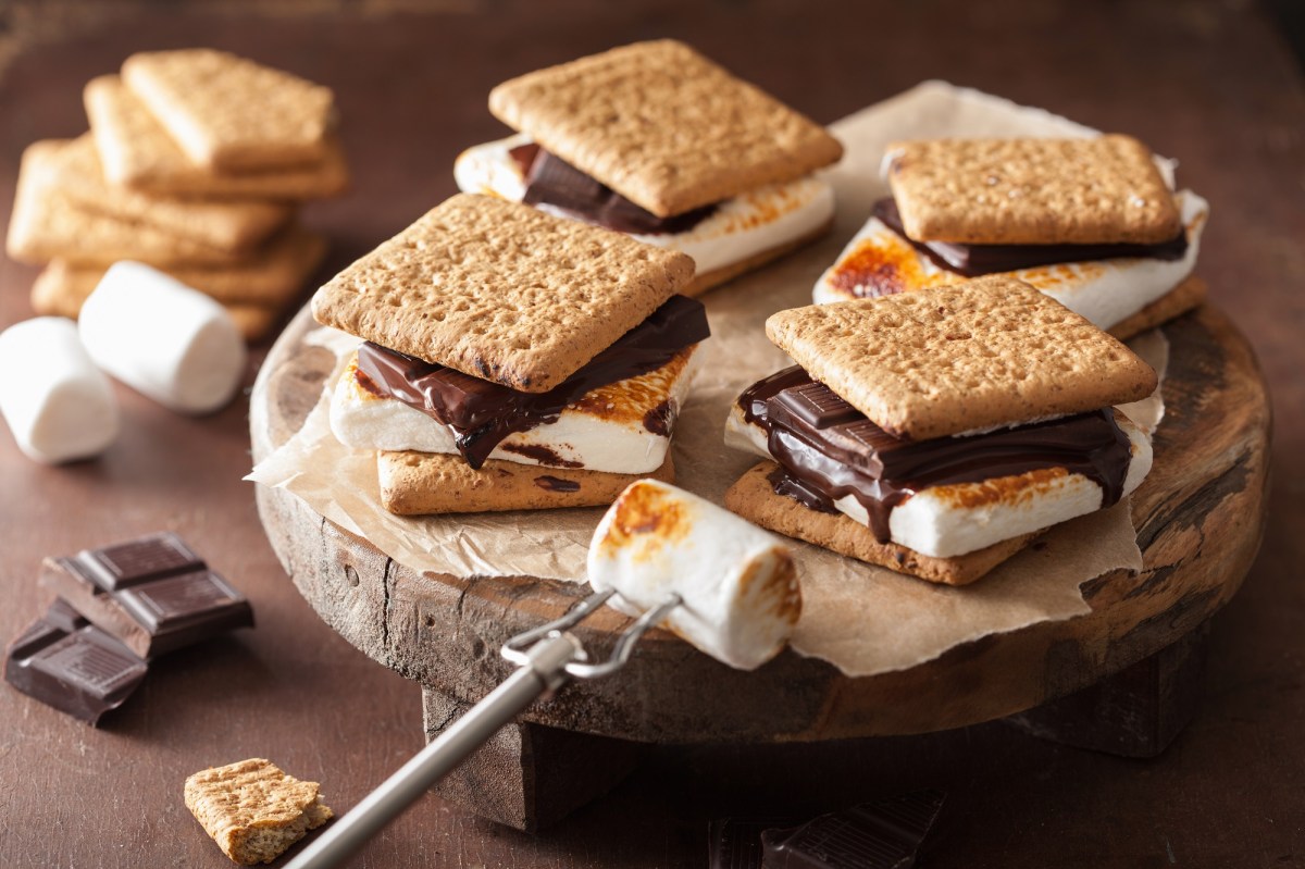 4 S'mores, kleine Sandwiches aus Keksen, gegrillten Marshmallows und Schokolade, auf einem runden Brett. Daneben die Zutaten.