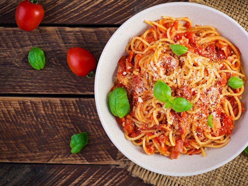 Ein Teller Spaghetti alla Amatriciana mit frischen Basilikumblättern in der Draufsicht. Daneben liegen Tomaten und Basilikumblätter.