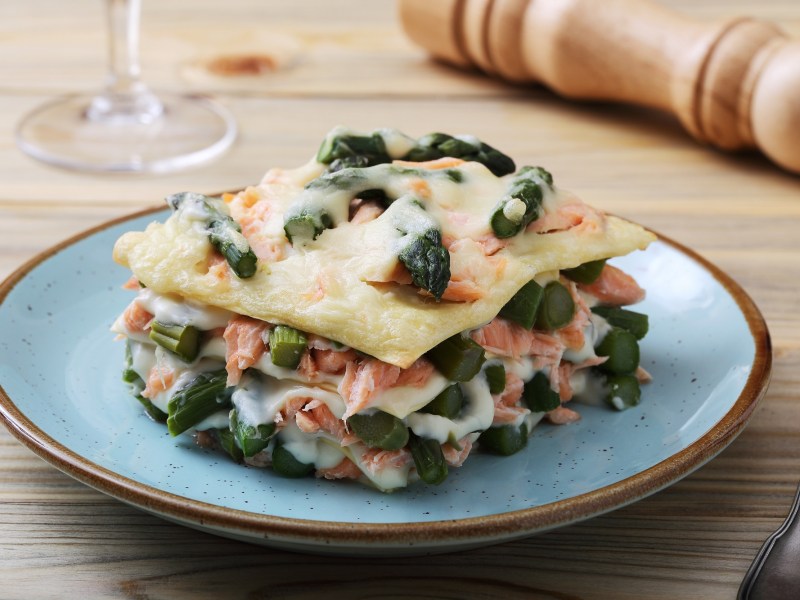 Ein Stück Spargel-Lachs-Lasagne auf einem hellblauen Teller. Daneben liegt eine Gabel und im Hintergrund liegt eine Pfeffermühle.