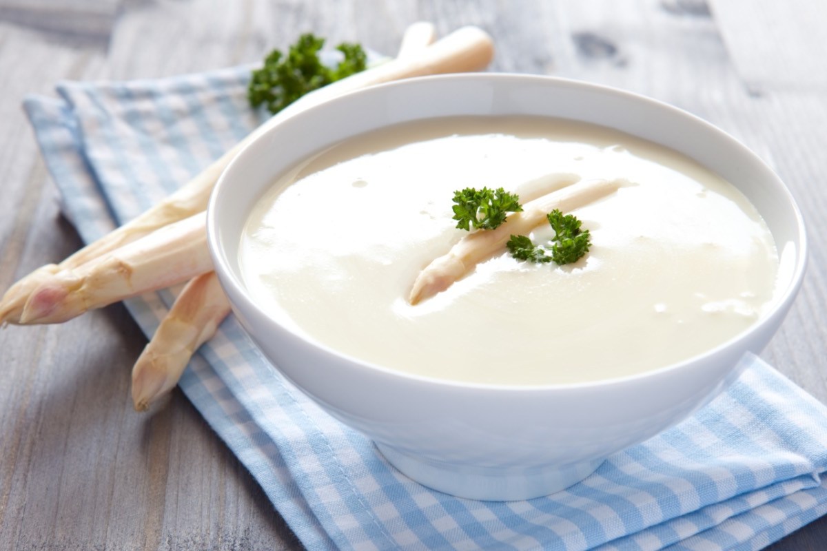 Eine Schüssel Spargelcremesuppe mit Petersilie. Frische Spargelstangen liegen daneben.