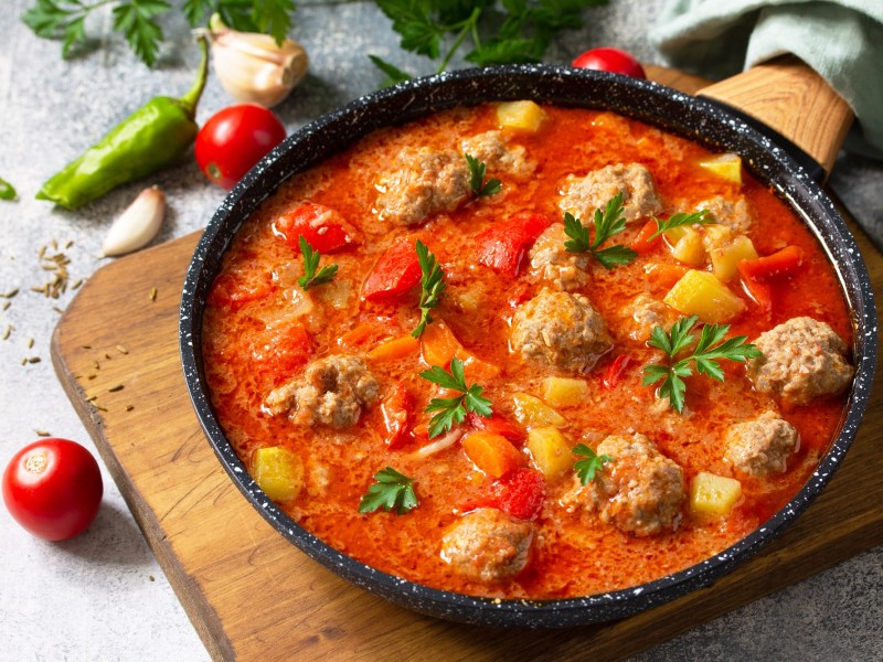 Tomatensuppe mit Hackbällchen in einer gusseisernen Pfanne, garniert mit Kräutern, daneben kleine Tomaten.