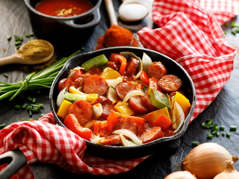 Würstchengulasch in einer gusseisernen Pfanne. Diese steht auf einem rot-weiß karierten Tischtusch. Daneben liegen Zwiebeln und Tomatenpulp.