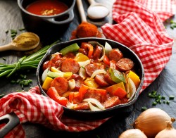 Würstchengulasch in einer gusseisernen Pfanne. Diese steht auf einem rot-weiß karierten Tischtusch. Daneben liegen Zwiebeln und Tomatenpulp.