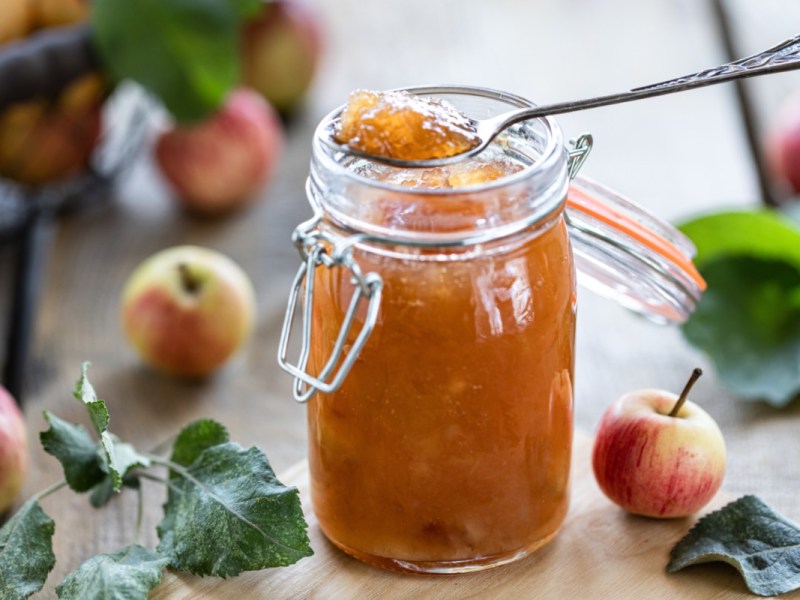 Ein Glas Apfelmarmelade mit Senf, in der ein Löffel steckt. Drumherum liegen Äpfel.