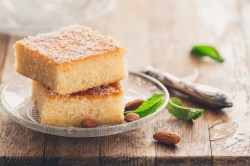 Basbousa, arabischer Gewürzkuchen auf einem Glasteller. Daneben liegen Mandeln und etwas Minze.