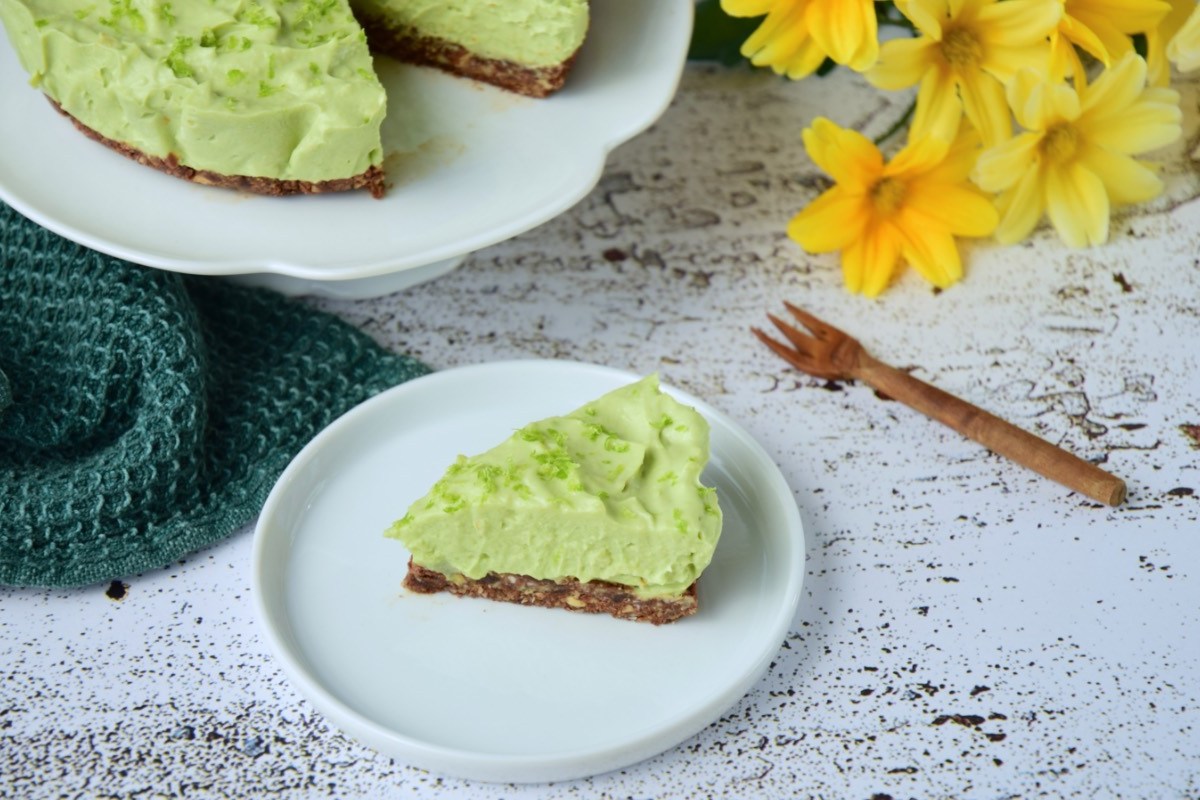 Ein Stück Avocado-Limetten-Käsekuchen auf einem Teller. Im Hintergrund der restliche Kuchen und ein paar gelbe Narzissen.