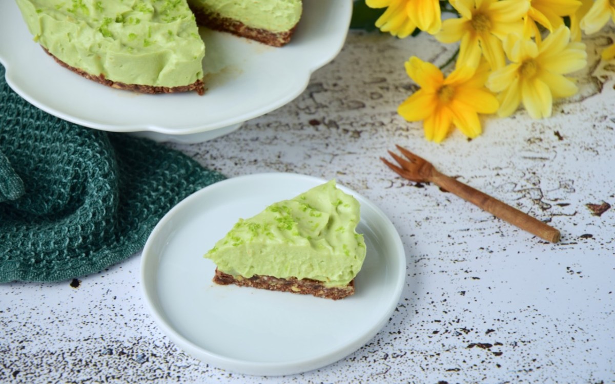 Ein Stück Avocado-Limetten-Käsekuchen auf einem Teller. Im Hintergrund der restliche Kuchen und ein paar gelbe Narzissen.