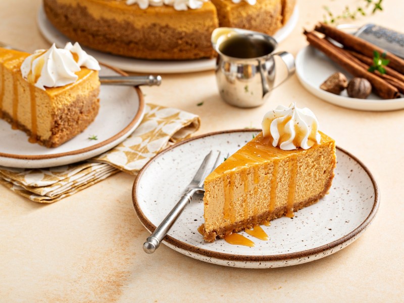 Chai Latte Cheesecake mit Karamellsoße und Sahne auf einem Teller. Im Hintergrund mehr Kuchen und Gewürze.