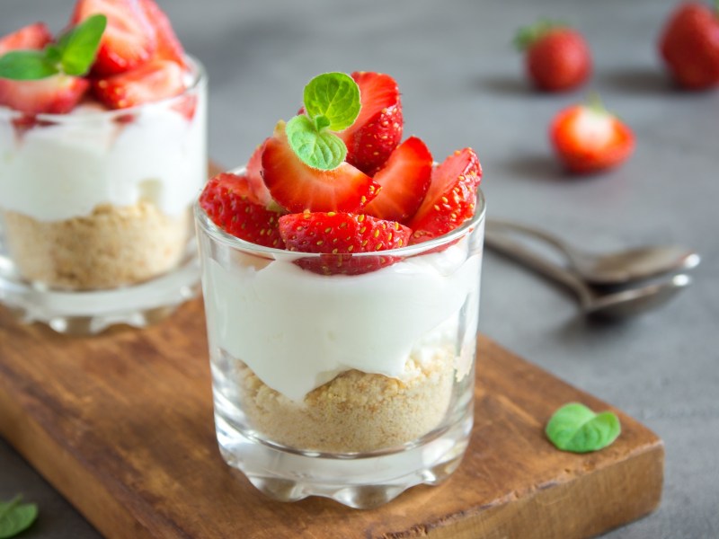 2 Cheesecake im Glas mit Keksboden und Erdbeeren garniert auf einem Brett, im Hintergrund Löffel und Erdbeeren.