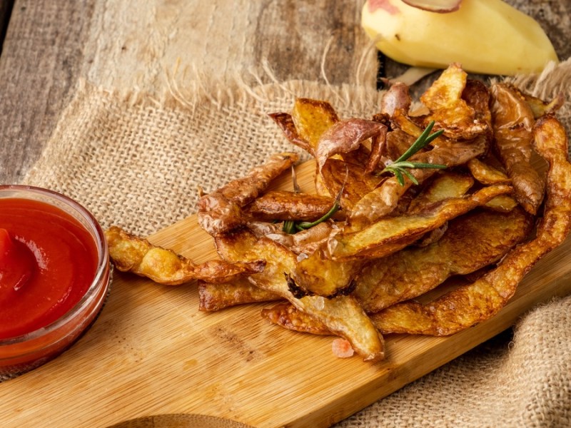 Chips aus Kartoffelschalen auf einem Holzbrett. Daneben eine Schale mit Ketchup.