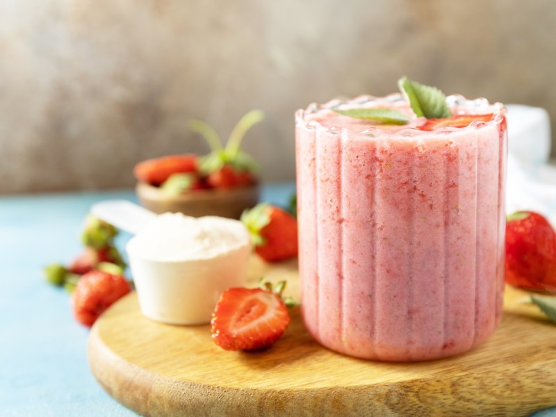 Ein Glas Erdbeer-Sahne-Shake mit Minze garniert auf einem Brett, daneben frische Beeren und Sahne in einer Schüssel.