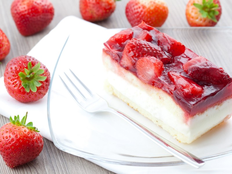 Ein Stück Erdbeer-Schmandkuchen vom Blech auf einem Teller, daneben eine Gabel und frische Erdbeeren.