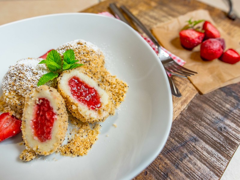 Ein weißer Teller mit Erdbeerknödel mit Semmelbrösel und ein Schneidebrett mit Erdbeeren daneben.