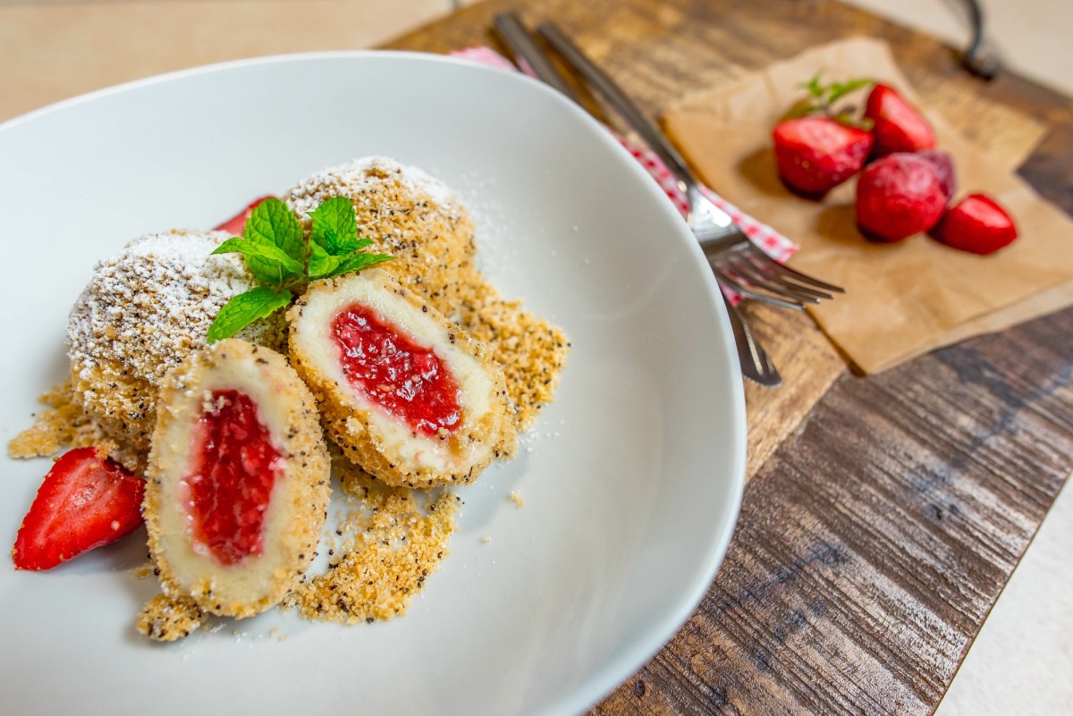 Ein weißer Teller mit Erdbeerknödel mit Semmelbrösel und ein Schneidebrett mit Erdbeeren daneben.