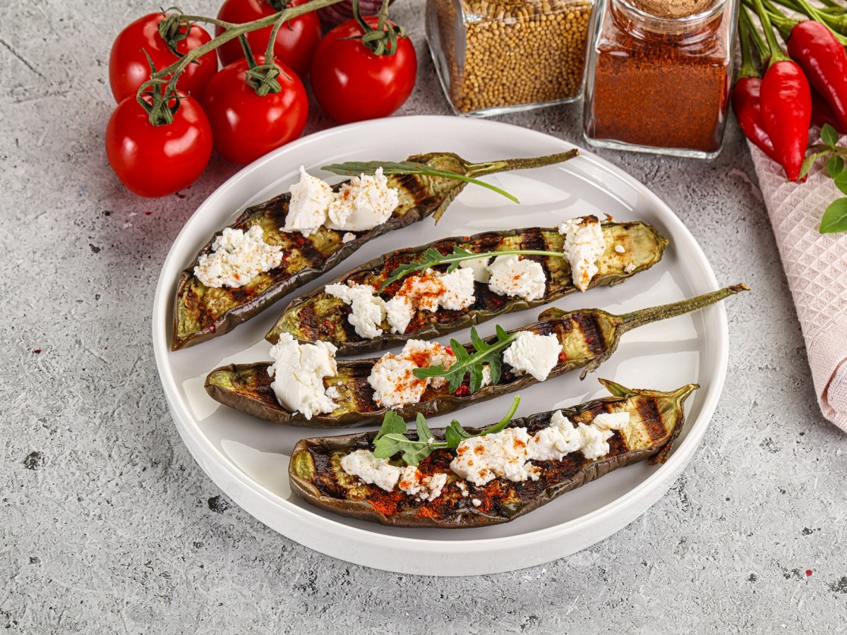 Gegrillte Aubergine mit Feta auf einem weißen Teller. Daneben liegen Kirschtomaten.