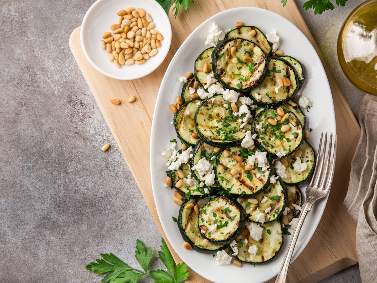 Gegrillte Zucchini in Scheiben mit Feta und Pinienkernen bestreut auf einem ovalen Teller, Draufsicht. Daneben Chiliflocken in einer Schüssel, eine Ölflasche und andere Zutaten.