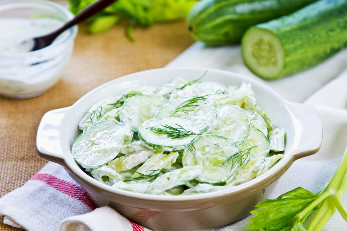 Eine Schüssel Gurkensalat mit Sahne, dahinter unscharf eine Schüssel mit der Salatsoße und Gurken.