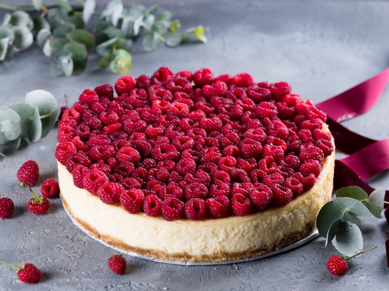 Himbeer-Kokos-Cheesecake dekoriert mit vielen Himbeeren, drumgerum ein paar Beeren und Zweige.