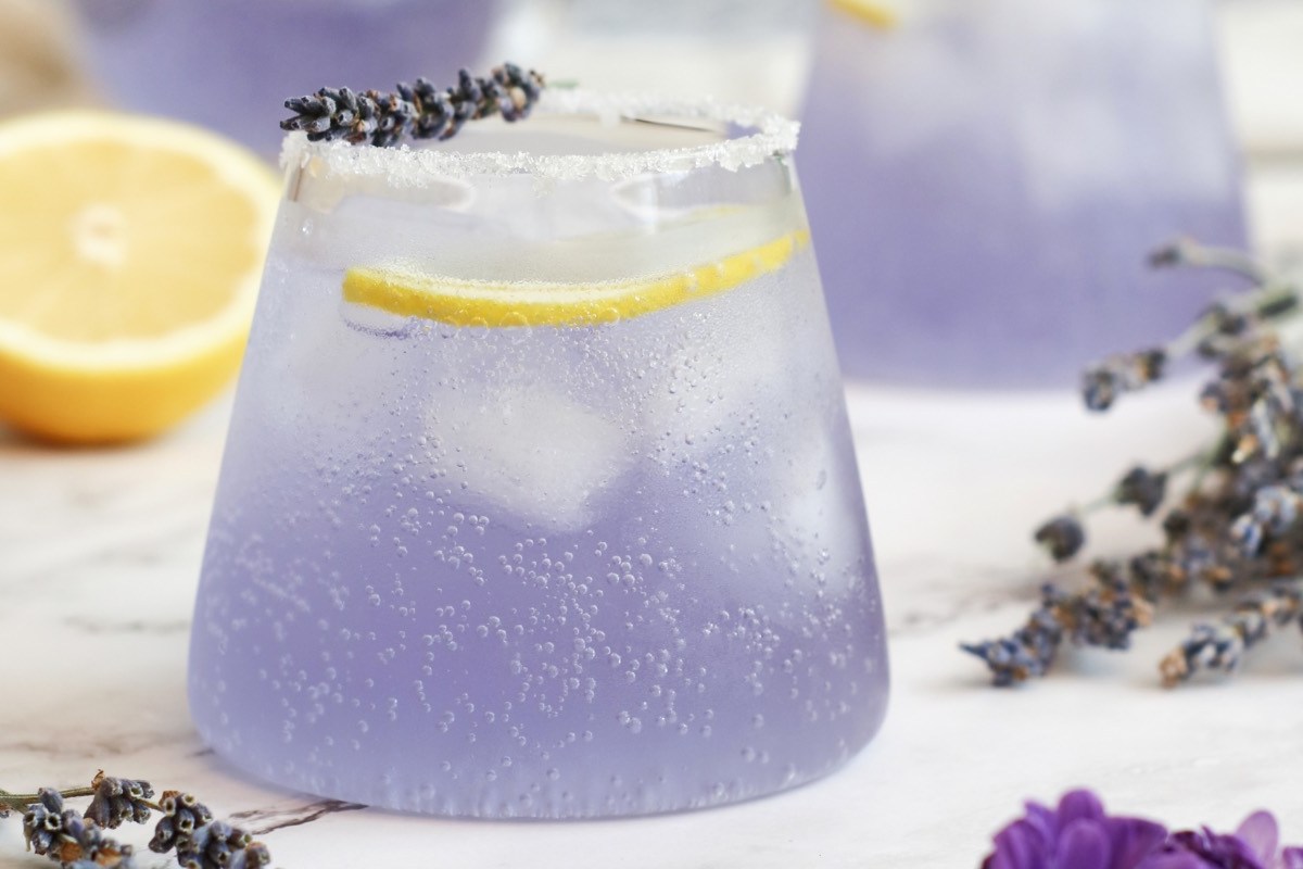 Ein Glas Holunder-Lavendel-Spritz. Drumherum liegen Lavendelblüten und eine halbe Zitrone.