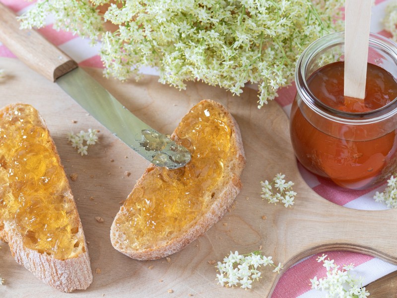 Zwei Brote mit Holunderblüten-Gelee, ein Glas Gelee und Holunderblüten daneben.