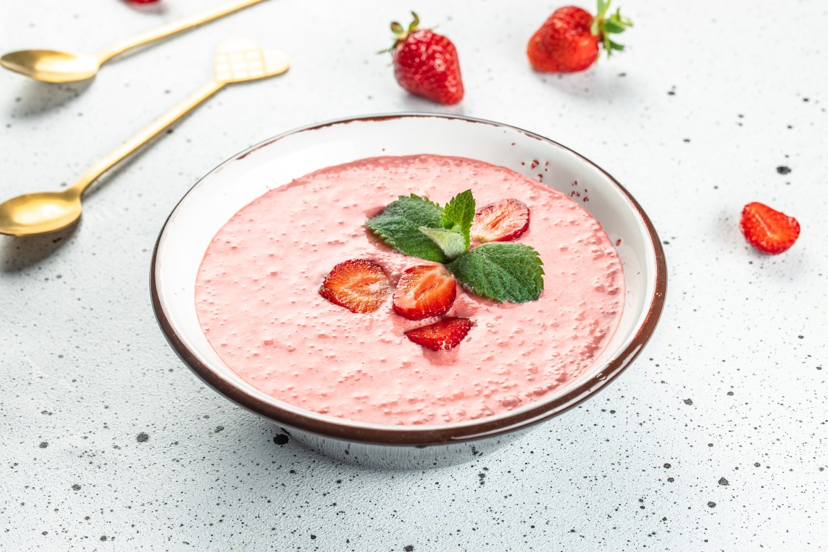 Eine Schüssel Erdbeersuppe garniert mit Erdbeeren und Minze, daneben ein paar Beeren und goldene Löffel.