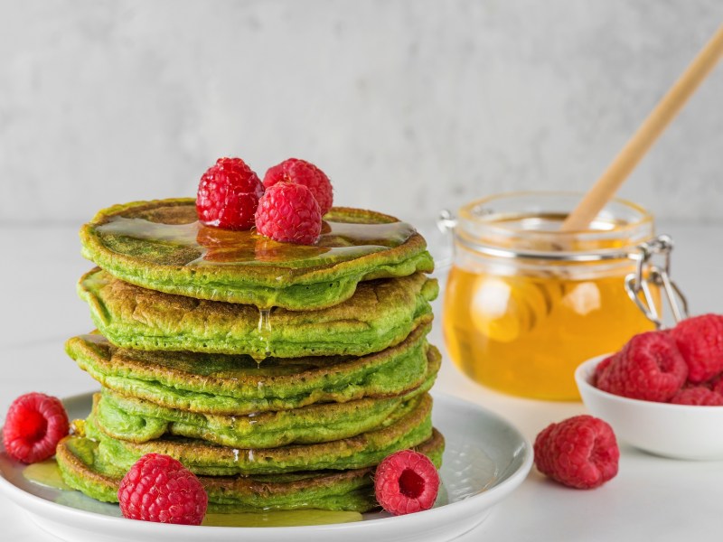 Ein Stapel Matcha-Pancakes. Garniert sind sie mit Honig und frischen Himbeeren.