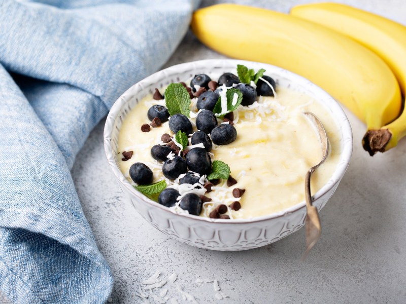 Nicecream in einer blauen Schüssel, garniert mit Beeren, Schokolade und Kokosraspeln. Daneben liegen Bananen