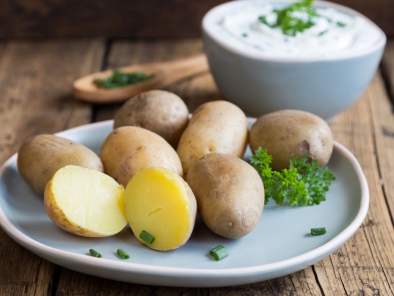 Pellkartoffeln auf einem Teller. Einige sind halbiert. Im Hintergrund steht eine Schüssel mit Kräuterquark.