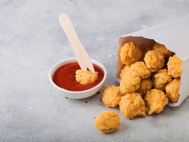 Popcorn Chicken in einer Papiertüte, aus der einige Stücke herauskullern. Daneben Ketchup