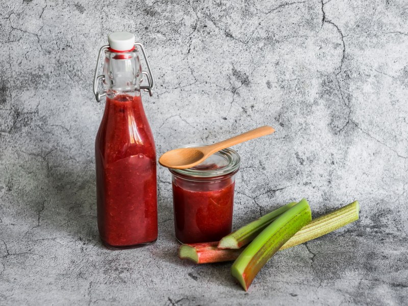 Flasche und Glas mit Rhabarber-Ketchup, Holzlöffel und Rhabarberstangen.