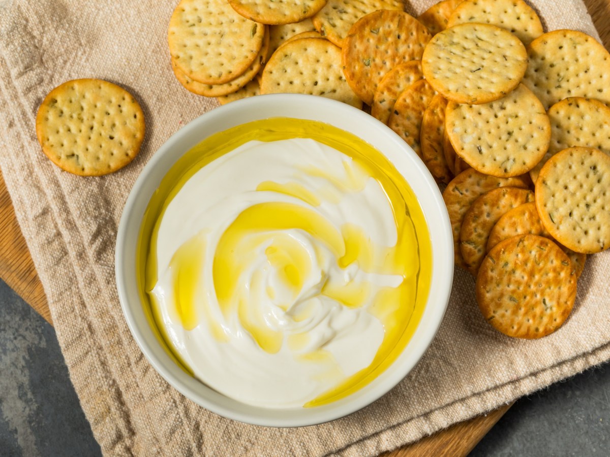 Ricotta-Dip mit Olivenöl neben einigen Crackern auf einem Holzbrett.